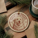 A circular ornament featuring a dog's face, with the text "GREAT PYRENEES" around the edge, is displayed on a wooden surface alongside a cup of coffee and some pine branches.