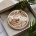 A circular ornament with a dog image and the text "GREAT PIRENES" on it, placed in a box with pine branches.