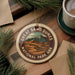 A circular ornament with the text "GREAT SAN DUNES NATIONAL PARK URK" is displayed on a wooden surface, surrounded by pine branches and a cup of coffee.