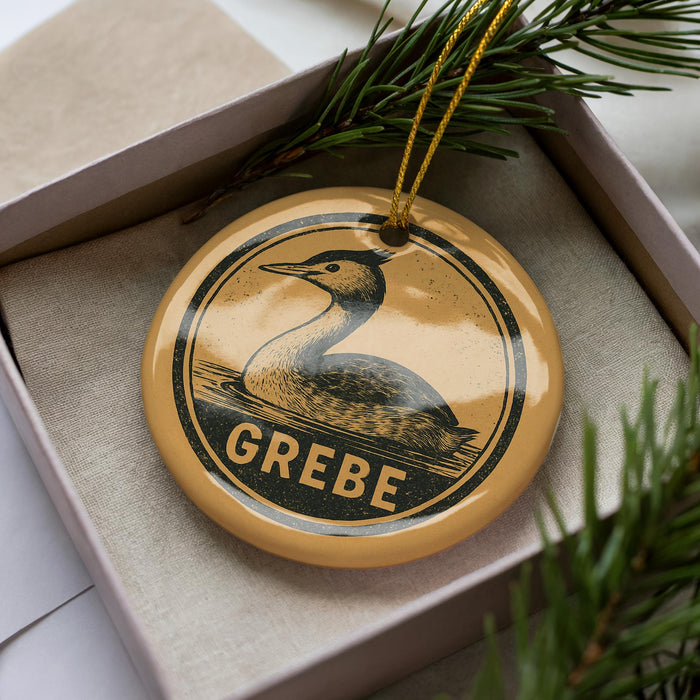 A circular ornament with a bird image and the word "GREBE" printed on it, placed inside a box with pine branches.