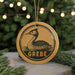 A circular ornament with a black and white illustration of a bird, possibly a crane, and the word "GREBE" written on it, hanging from a string.