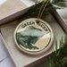 A circular ornament with a green iguana image and the text "GREEN GUANAN" on it, placed in a box with pine branches.