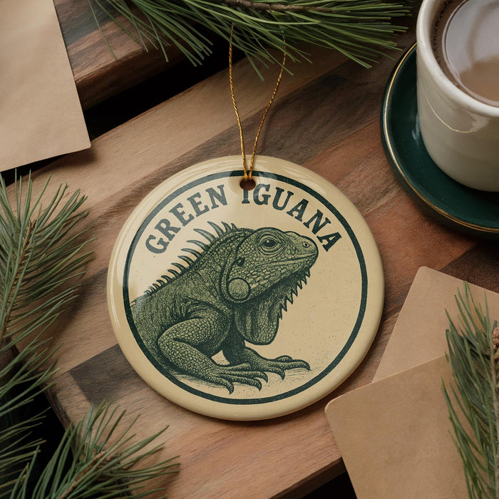 A circular ornament featuring a green iguana, with the text "GREEN GUANA" printed on it, is displayed on a wooden surface alongside a cup of coffee and some pine branches.