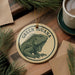 A circular ornament featuring a green iguana, with the text "GREEN GUANA" printed on it, is displayed on a wooden surface alongside a cup of coffee and some pine branches.