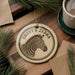 A circular ornament featuring a zebra's head is displayed on a wooden surface, surrounded by pine branches and a cup of coffee.