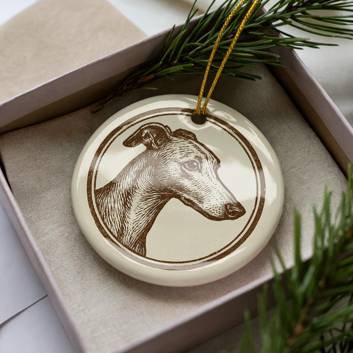 A ceramic ornament in the shape of a dog's head, featuring a brown and white design, is displayed in a box with pine branches.