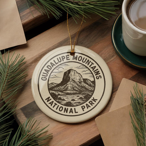 A ceramic ornament with the text "GUADALUPE MOUNTAINS NATIONAL PARK" and a mountain illustration is displayed on a wooden surface, accompanied by pine branches and a cup of coffee.
