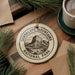 A ceramic ornament with the text "GUADALUPE MOUNTAINS NATIONAL PARK" and a mountain illustration is displayed on a wooden surface, accompanied by pine branches and a cup of coffee.