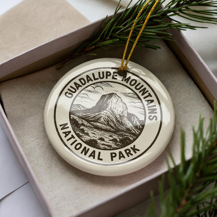 A white ornament with a black and white illustration of a mountain and the text "GUADALUPE MOUNTAINS NATIONAL PARK" is displayed in a box, surrounded by pine branches.