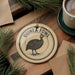 A decorative ornament featuring a guinea fowl, a type of bird, is displayed on a wooden surface alongside a cup of coffee and some pine branches.