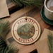 A circular ornament with the text "HALLSTAT" and "AUSTRIA" is displayed on a wooden surface, surrounded by pine branches and a cup of coffee.