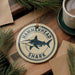 A round ornament with the text "HAMMERHEAD SHARK" and a graphic of a hammerhead shark is displayed on a wooden surface, accompanied by pine branches and a cup of coffee.