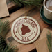 A circular ornament with the text "HAWAII'I ISLAND USA" printed on it, surrounded by pine branches and a cup of coffee on a wooden surface.