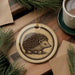 a round ornament with a porcupine design, placed on a wooden surface next to a cup of coffee and some pine branches.