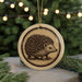 A round ornament featuring a porcupine design is displayed in front of a blurred background of Christmas lights.