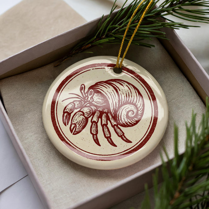 A round ornament with a red and white design depicting a hermit crab is displayed in a box, surrounded by pine branches.
