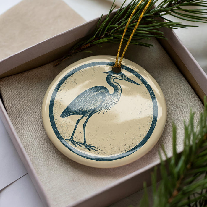 A ceramic ornament in the shape of a heron, with a blue and white design, is displayed in a box with pine branches.