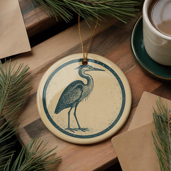 a round ornament with a blue and white design depicting a heron, placed on a wooden surface next to a cup of coffee and some pine branches.