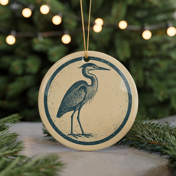 A round ornament featuring a blue heron is displayed against a blurred background of a Christmas tree with twinkling lights.