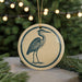 A round ornament featuring a blue heron is displayed against a blurred background of a Christmas tree with twinkling lights.