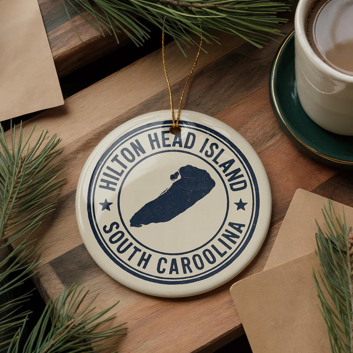 A ceramic ornament with the text "Hilton Head Island South Carolina" printed on it, surrounded by pine branches and a cup of coffee on a wooden surface.