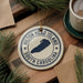 A ceramic ornament with the text "Hilton Head Island South Carolina" printed on it, surrounded by pine branches and a cup of coffee on a wooden surface.