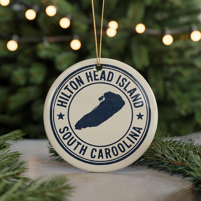 A white ornament with a blue design, featuring the text "Hilton Head Island South Carolina" and a map of the island, is displayed on a Christmas tree with twinkling lights in the background.