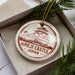 A circular ornament with a red and white design, featuring a castle-like structure, is displayed in a box with pine branches.