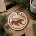 A round ornament featuring a brown badger, with the text "HONEY BADGER" printed on it, is displayed on a wooden surface alongside a cup of coffee and some pine branches.