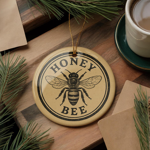 A round ornament with the text "HONEY BEE" and a bee illustration is displayed on a wooden surface, surrounded by pine branches and a cup of coffee.