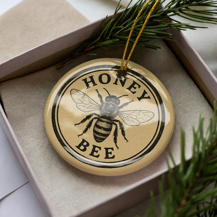 A round ornament with the text "HONEY BEER" and a bee illustration is displayed in a box, surrounded by pine branches.