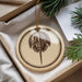 A circular ornament with a brown design featuring a stylized crab or lobster is displayed in a box, surrounded by pine branches.
