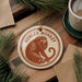 A round ornament featuring a monkey sitting on a branch, with the text "HOWLER MONKEY" printed on it. The ornament is hanging from a string, and there is a cup of coffee and some pine branches in the background.