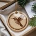 A round ornament with a brown bird design is displayed in a box, surrounded by pine branches and greenery.