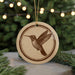 A round ornament featuring a hummingbird is displayed in front of a blurred background of Christmas lights.