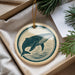 A round ornament featuring a whale design is displayed in a box, surrounded by pine branches.