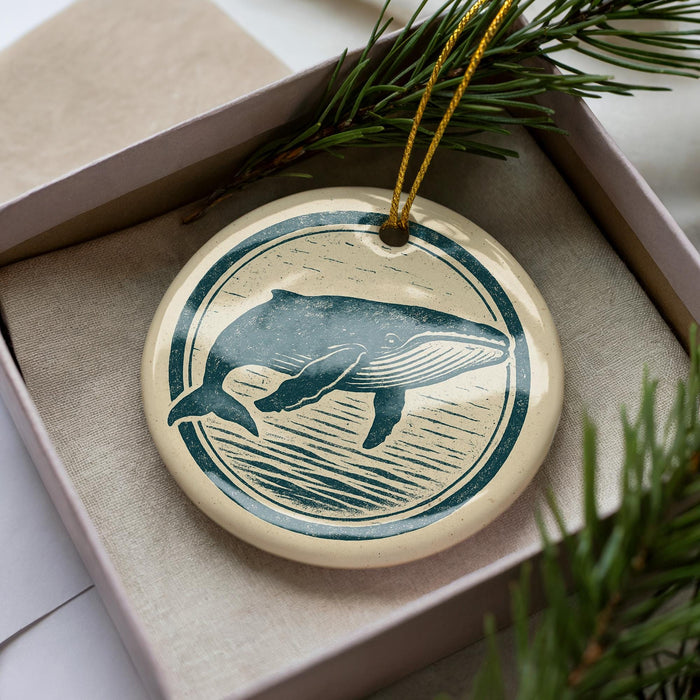 A ceramic ornament in the shape of a whale, with a circular design featuring a silhouette of the whale and a wave pattern, is displayed in a box with pine branches.