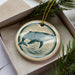 A ceramic ornament in the shape of a whale, with a circular design featuring a silhouette of the whale and a wave pattern, is displayed in a box with pine branches.