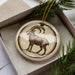A decorative ornament featuring a brown goat with a curved horn, displayed in a box with pine branches.