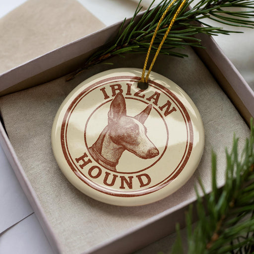 A circular ornament with the text "IBAN HOUND" printed on it, featuring a hound's head image. The ornament is placed inside a box, surrounded by pine branches.