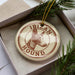 A circular ornament with the text "IBAN HOUND" printed on it, featuring a hound's head image. The ornament is placed inside a box, surrounded by pine branches.
