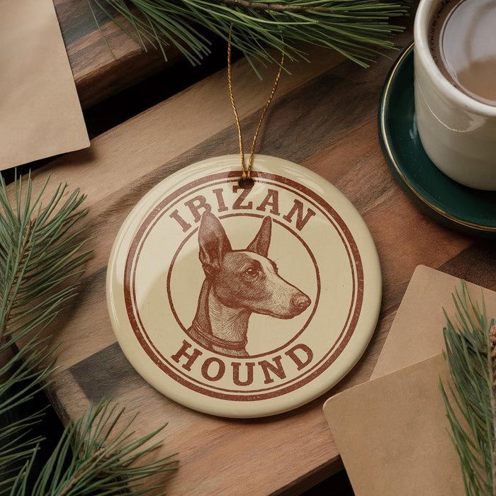 A circular ornament with the text "IBIZAN HOUND" and an illustration of a dog's head is displayed on a wooden surface, surrounded by pine branches and a cup of coffee.