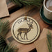 A circular ornament featuring an illustration of an impala deer, with the word "IMPALA" written on it, is displayed on a wooden surface alongside a cup of coffee and some pine branches.