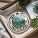 A decorative ornament with a mountain landscape and Swiss flag design, placed in a box with pine branches.
