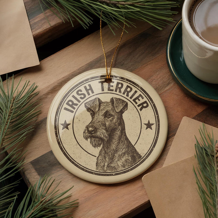 A circular ornament featuring an illustration of an Irish Terrier dog, with the text "IRISH TERRIER" prominently displayed. The ornament is hanging from a string, and there are pine branches and a cup of coffee visible in the background.
