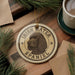 A circular ornament featuring an image of an Irish Water Spaniel dog, surrounded by pine branches and a cup of coffee on a wooden surface.