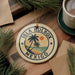 A circular ornament with the text "ISLA HOLBOX MEXICO" and a palm tree design, hanging on a wooden surface next to a cup of coffee and some pine branches.