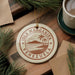 A circular ornament with the text "ISLA MARGARITA VENEZUELA" printed on it, surrounded by pine branches and a cup of coffee on a wooden surface.