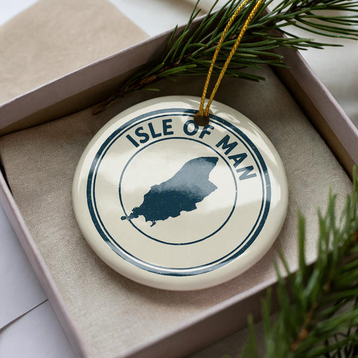 A white ornament with a blue circular design featuring the silhouette of the Isle of Man, surrounded by pine branches and a yellow string.