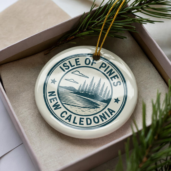 A white ornament with a blue design, featuring the text "ISLE OF PINES NEW CALEDONIA" and a landscape with pine trees and a body of water.